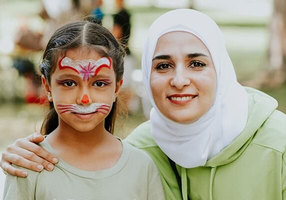 A child wears facepaint and smiles for a photo with a woman next to her